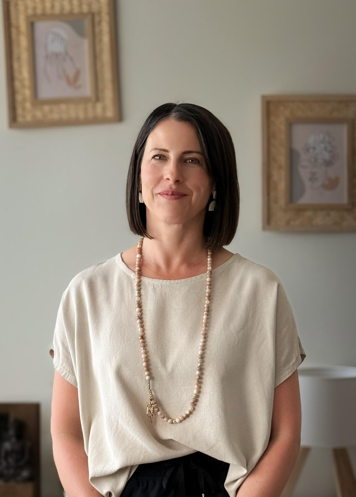 Gemma reflexology therapist Woman wearing cream top, black pants and a mala bead necklace stood in a room smiling.